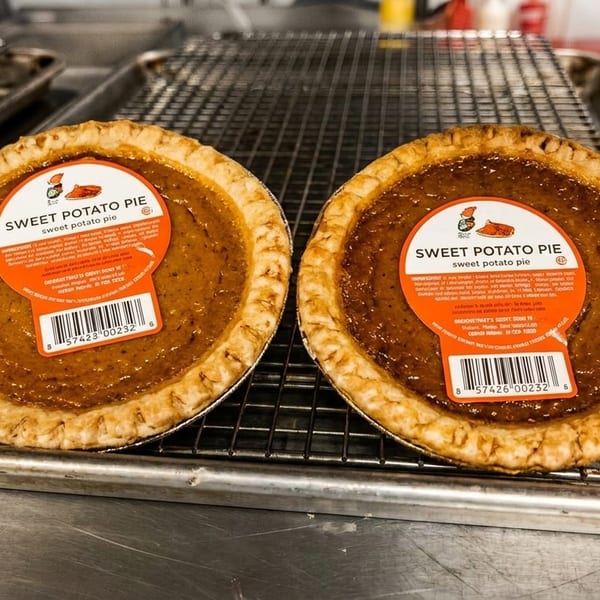 Two sweet potato pies with orange labels sit on metal cooling racks in a commercial kitchen.